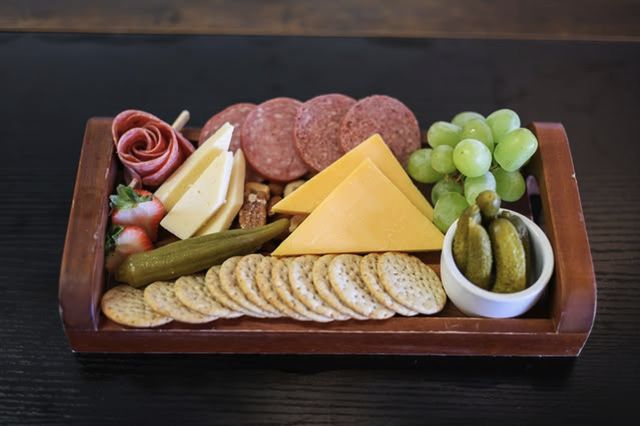 A wooden tray of cheeses, salami, grapes, pickles, and crackers